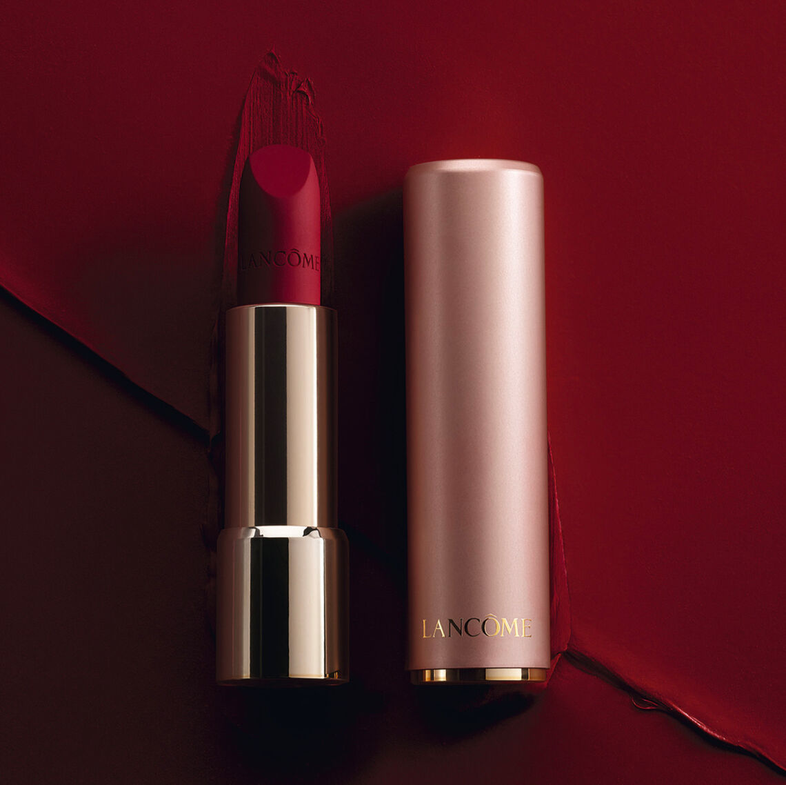 A lipstick in a sleek silver and gold tube, placed beside its matching cylindrical case, against a deep red background with a smear of lipstick.