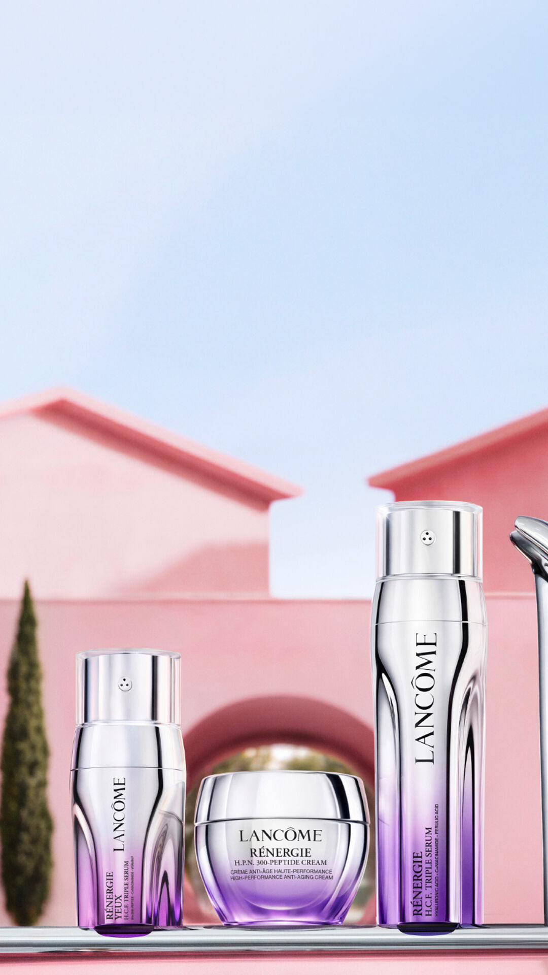 Renergie skincareproducten, waaronder een oogcreme, anti-aging creme en serum, gepresenteerd op een glanzend roze oppervlak voor een roze villa met cipressen en een blauwe hemel.