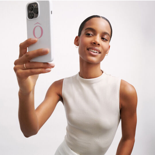 Een vrouw met een gladde huid en een strakke, lichte top houdt een smartphone vast en kijkt lachend naar de camera.