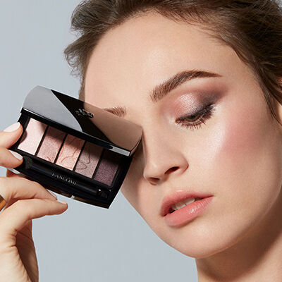 A woman applying eyeshadow while holding a sleek eyeshadow palette near her face, showcasing a soft, natural makeup look.