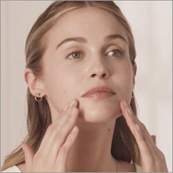 A young woman gently applying skincare to her face, looking upward with a thoughtful expression.