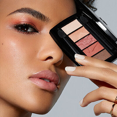 A close-up of a person holding a compact eyeshadow palette near their face, showcasing shimmery eyes and a subtle lip look against a light background.