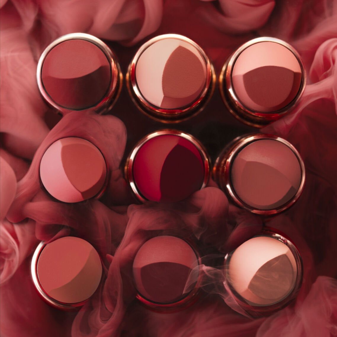 A flat lay of nine round containers of varying shades of reddish lip products, surrounded by soft, swirling smoke.
