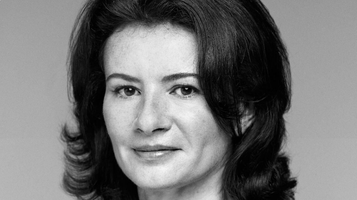 Portrait en noir et blanc d'une femme souriante, avec des cheveux bruns et ondules, fond uni.