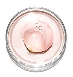 A round clear container holds a soft pink gel, featuring a delicate rose petal and a few droplets of water on its surface.