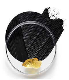 A transparent petri dish containing a piece of gold sits in front of a textured black background with diagonal brush strokes.