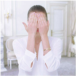 A woman in a white blouse covers her face with her hands, standing in an elegantly decorated room.
