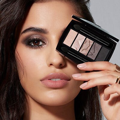 A model holds a compact eyeshadow palette close to her face, showcasing smoky eye makeup and neutral lips.