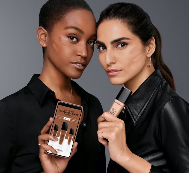 Two women of diverse backgrounds pose together, holding makeup products while showcasing a smartphone displaying cosmetics. The background is a solid gray.
