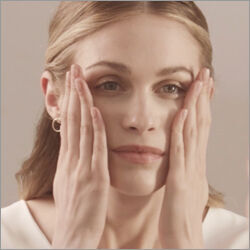 A woman with light brown hair gently massages her face with both hands, expressing a calm and relaxed demeanor. She is wearing subtle makeup and gold hoop earrings against a neutral background.