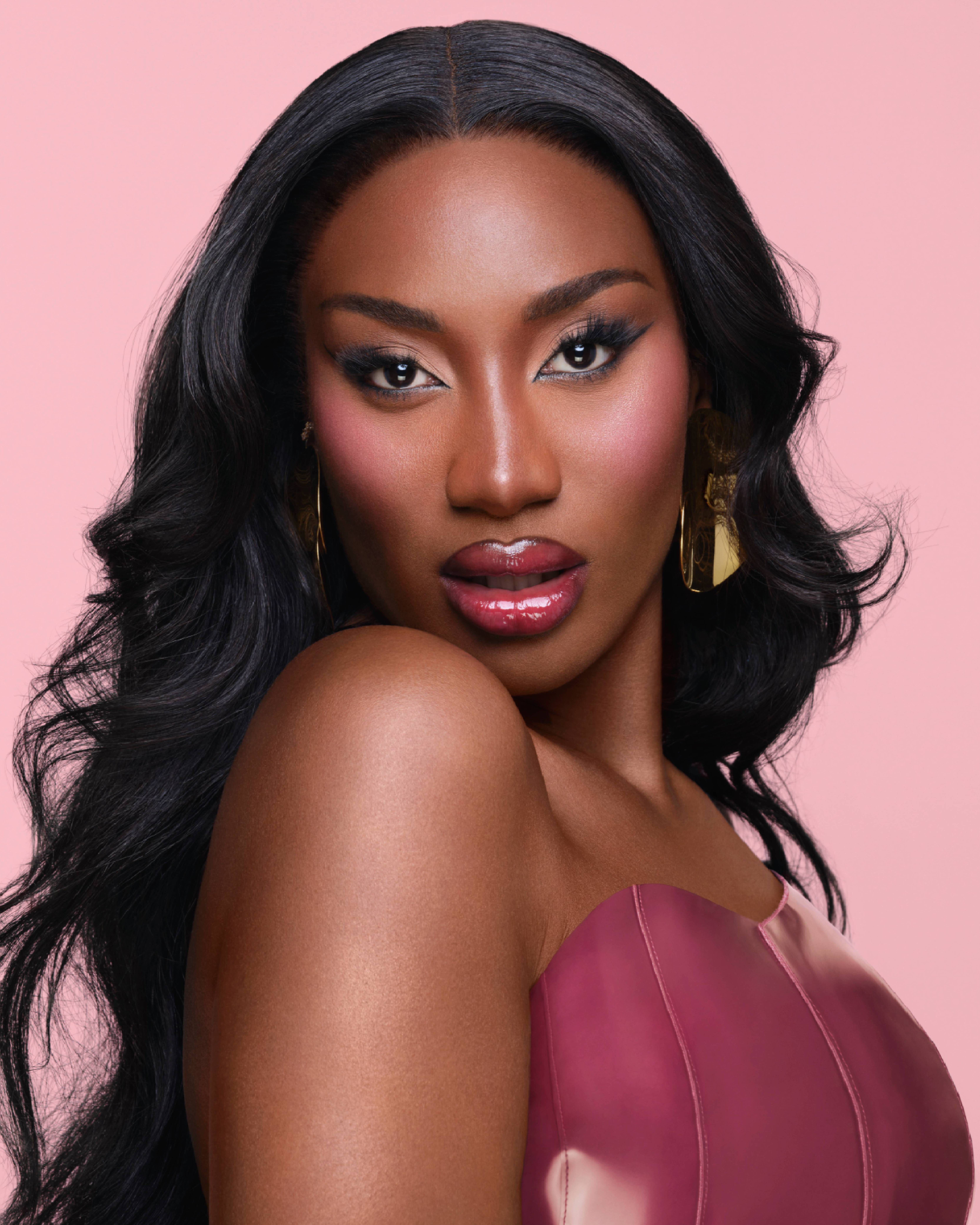 Portrait d'une femme avec des cheveux longs et noirs, portant un maquillage audacieux et des boucles d'oreilles dorees, sur fond rose.