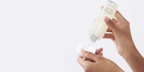 Une personne verse un liquide d'une bouteille sur un coton, sur un fond clair.