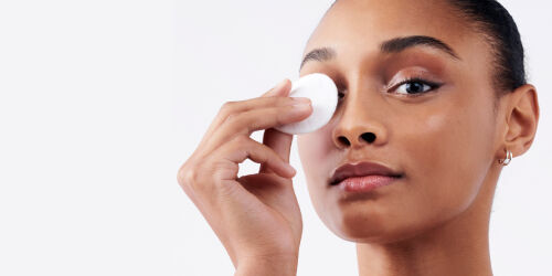 Femme appliquant un coton sur son oeil, avec une peau eclatante, sur fond blanc.