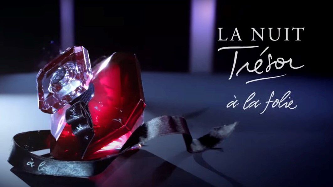 Flacon de parfum La Nuit Tresor a la folie, rouge et transparent, pose sur une surface sombre avec un ruban noir.